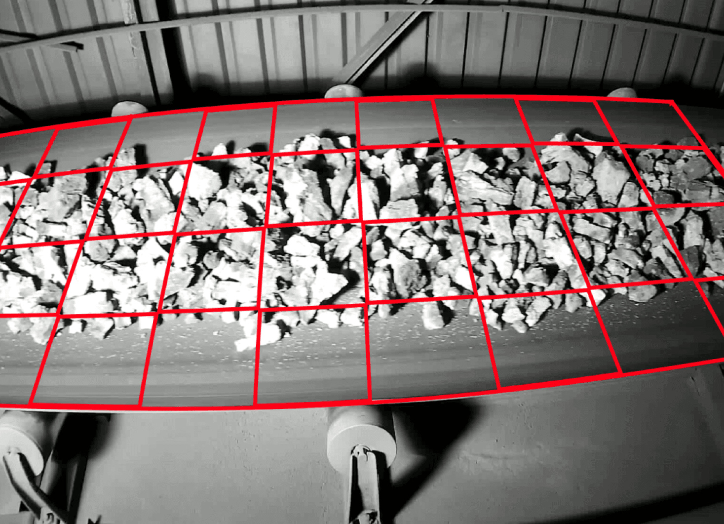 Conveyor belt with scattered rocks, overlaid with a red grid, inside an industrial setting.
